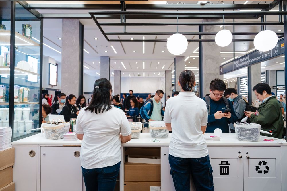 Modern retail store interior with customers browsing products at white display counters and pendant lighting.