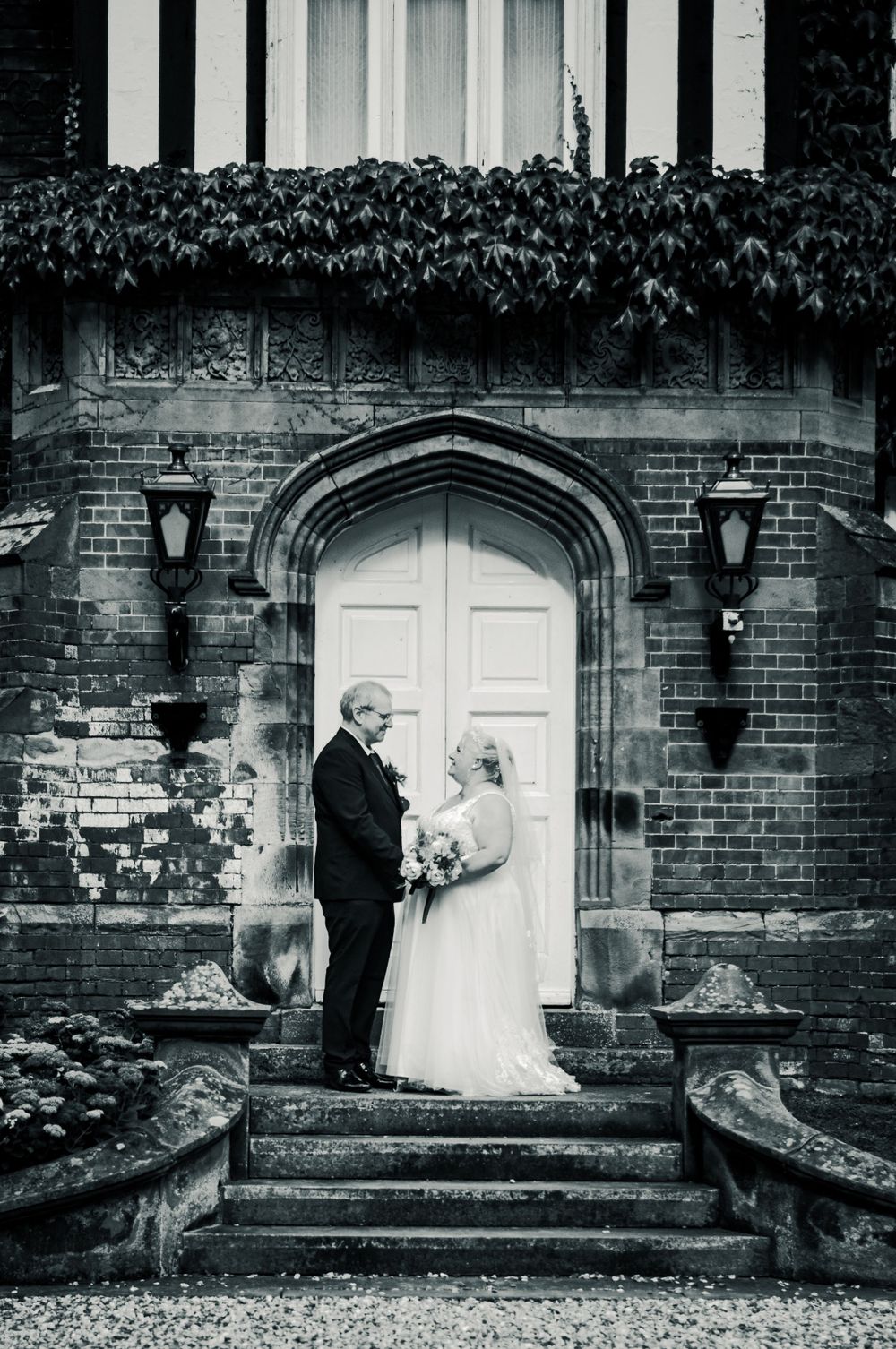 Classic black and white wedding portrait on steps of ivy-covered brick building.