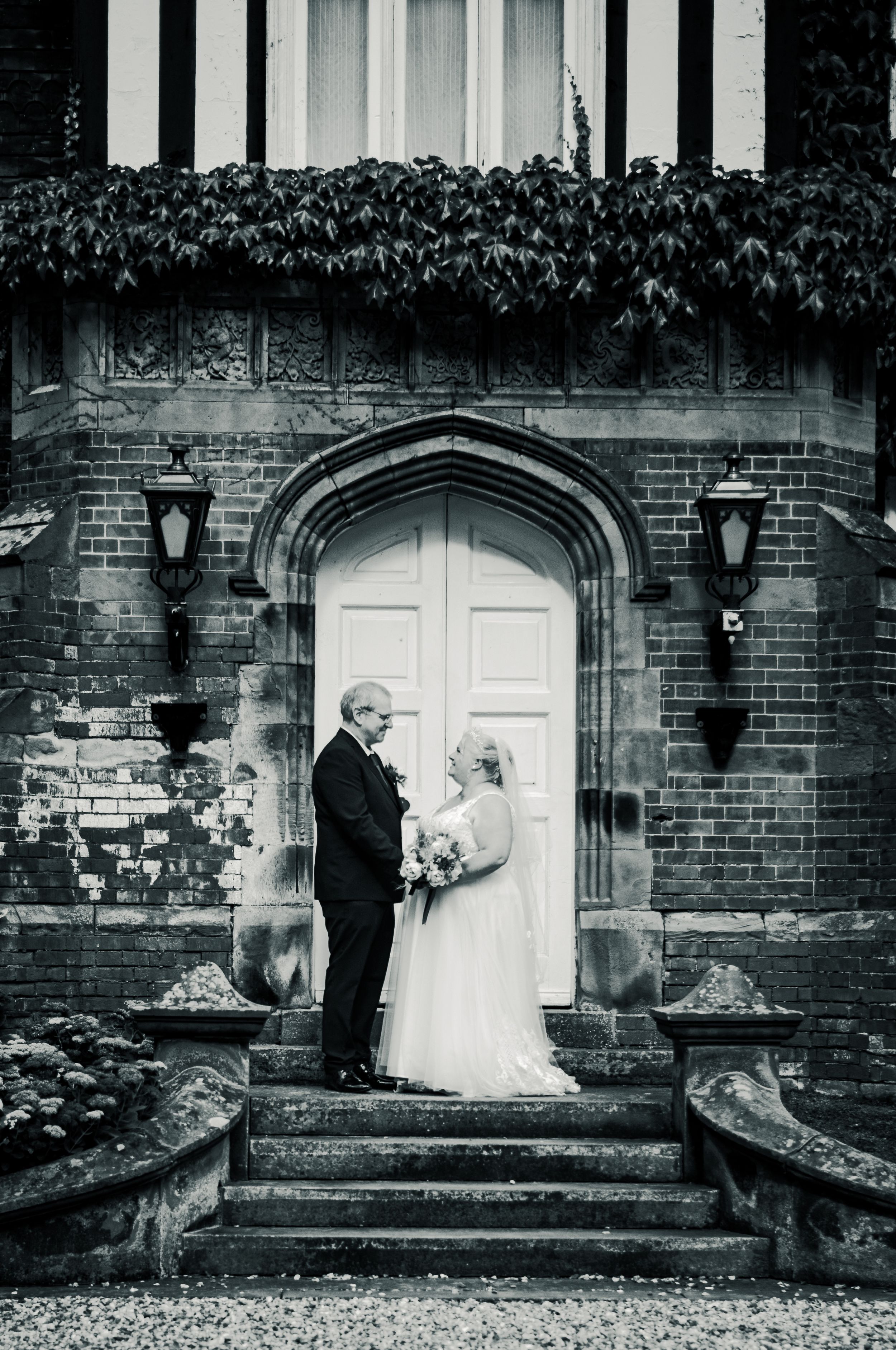 Classic black and white wedding portrait on steps of ivy-covered brick building.