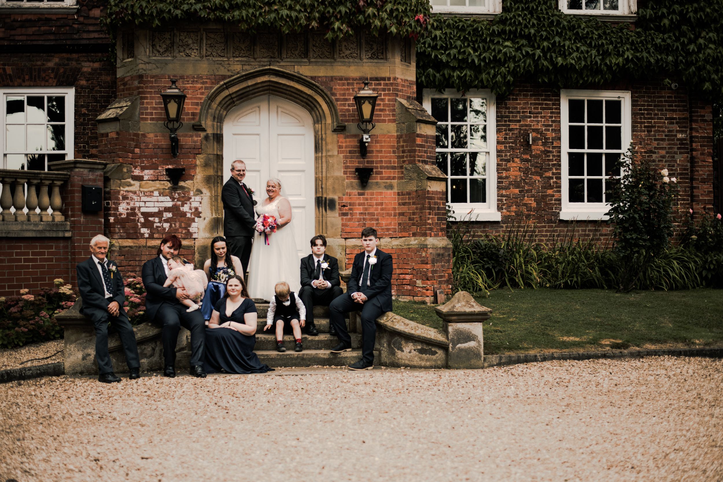Wedding party poses on stone benches in front of historic brick building with ivy.