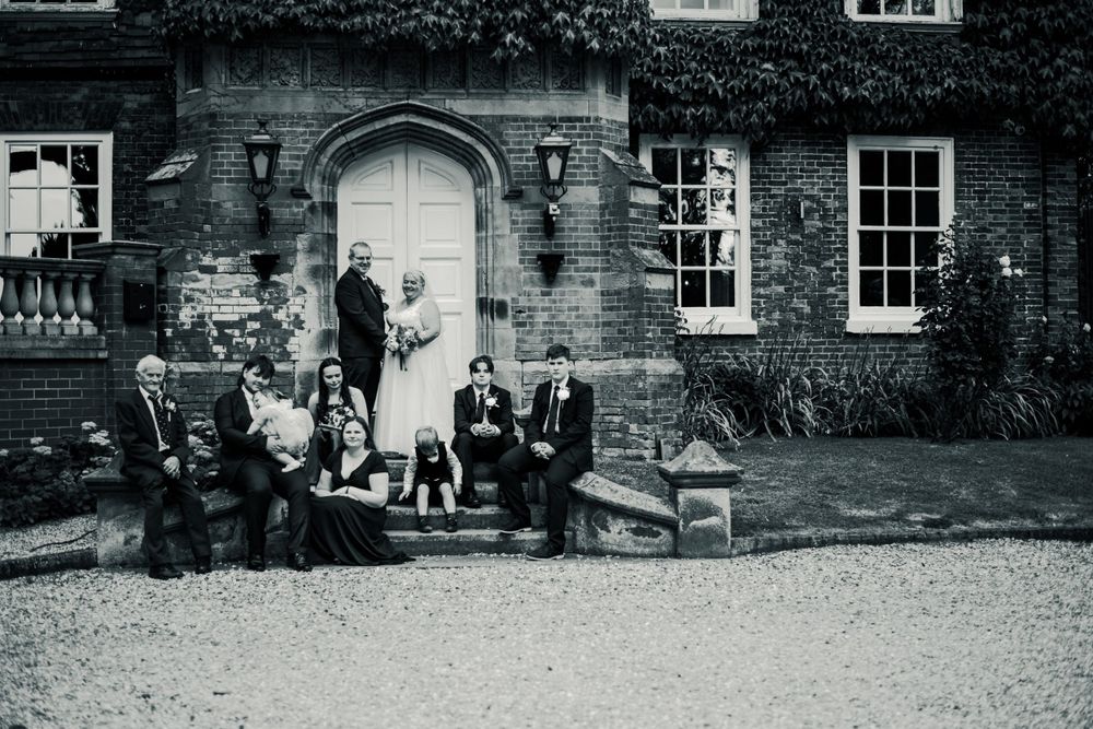 Black and white photo of wedding party gathered on stone benches by historic doorway.
