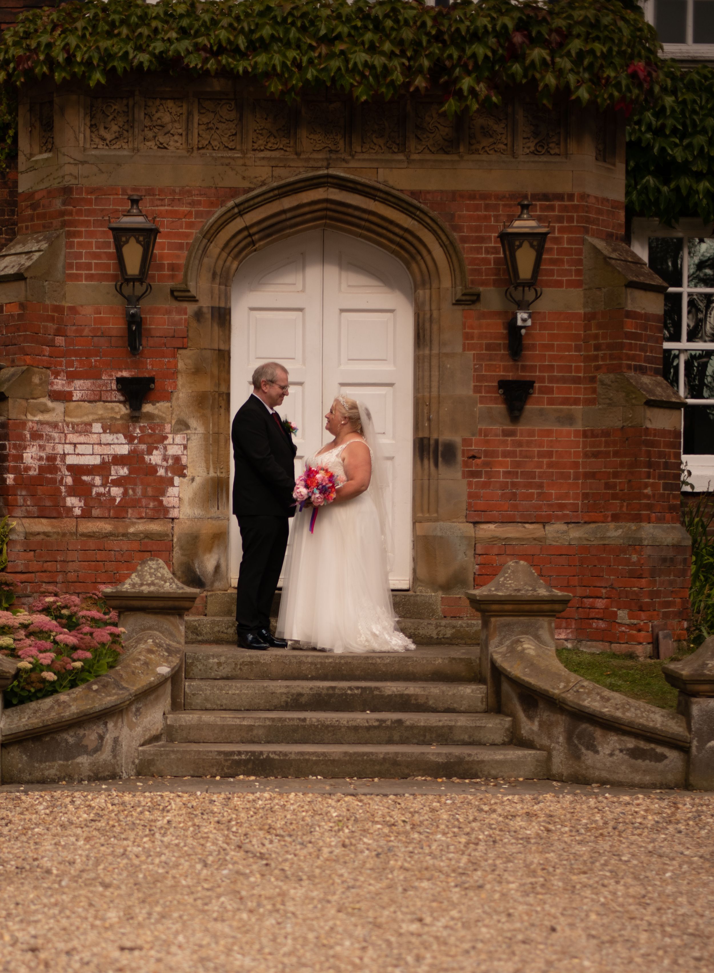 Couple stands on stone steps before arched doorway of ivy-covered brick building.