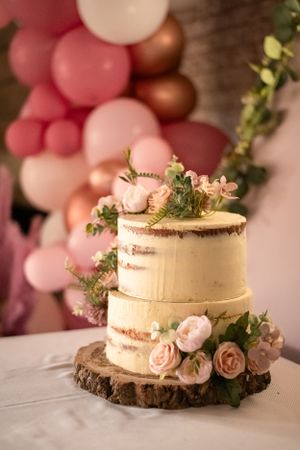 Elegant two-tier semi-naked wedding cake decorated with pink roses on wood slice with balloon backdrop.