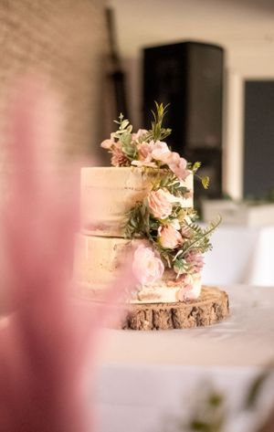 Semi-naked wedding cake decorated with fresh pink flowers on rustic wood slice stand.