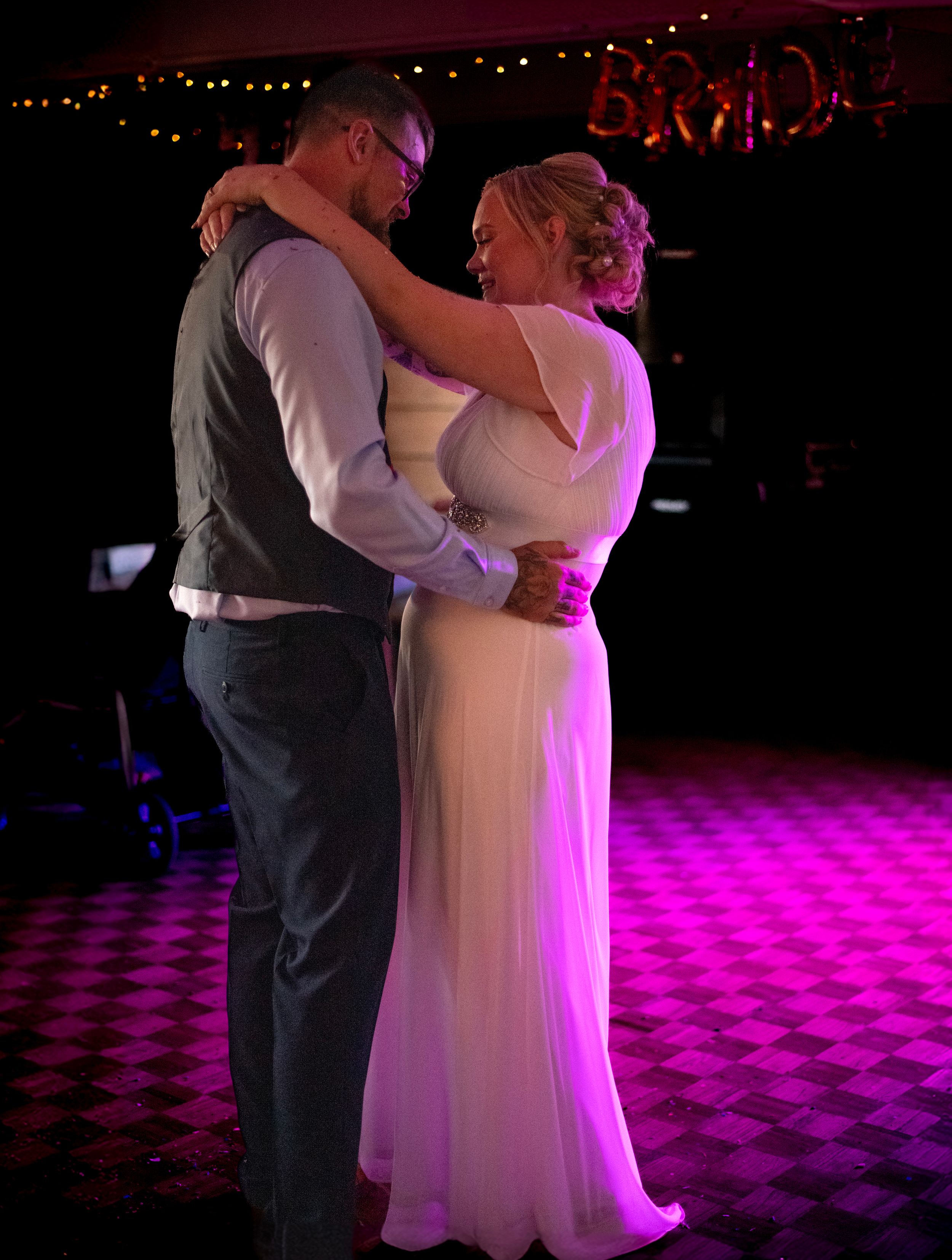 A romantic first dance moment on a checkered floor under pink lighting at a wedding reception.