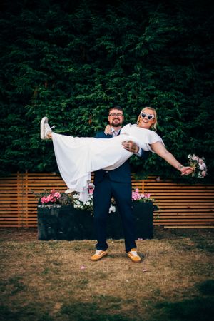 A couple shares a playful moment during their wedding as the groom lifts the bride against a backdrop of greenery.