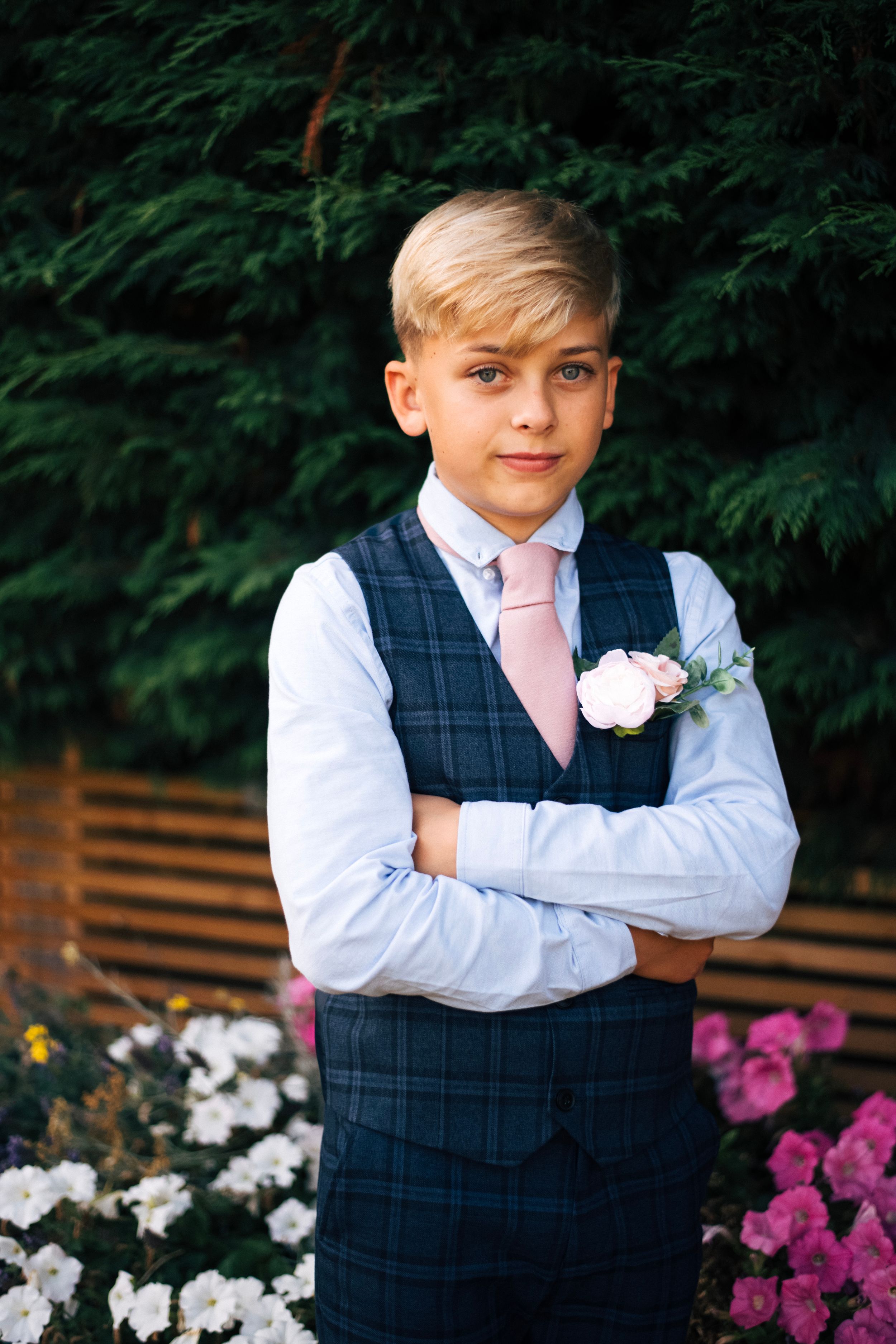 Young attendant in a navy checkered vest and pink tie poses formally in front of garden flowers.