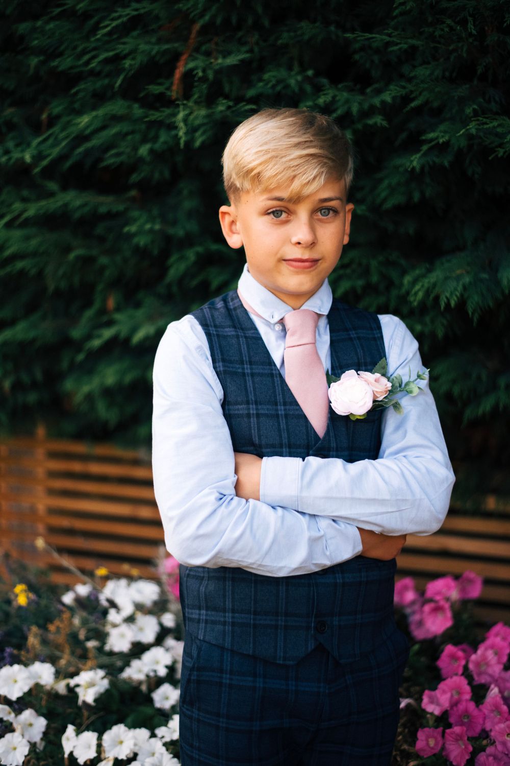 Young attendant in a navy checkered vest and pink tie poses formally in front of garden flowers.