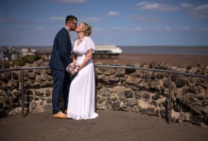 Romantic wedding couple embracing on stone overlook with coastal view in background.