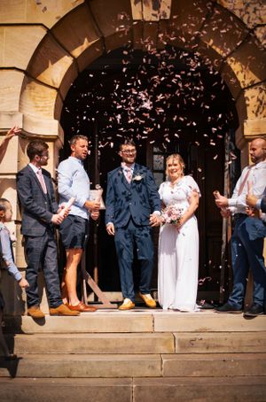 Wedding party celebration with confetti shower on church steps under stone archway.