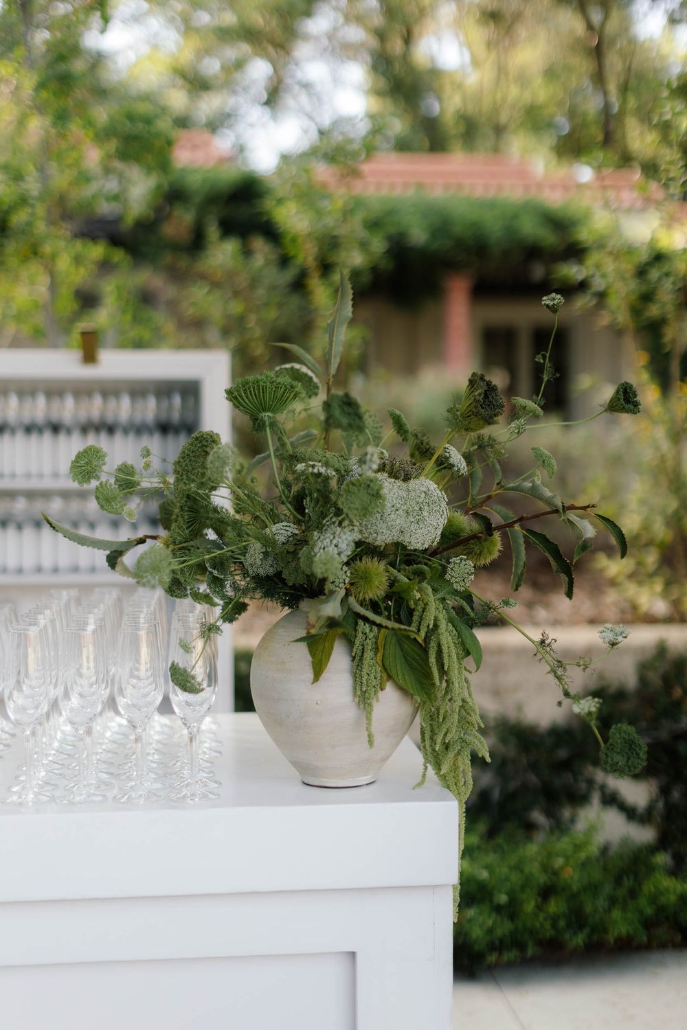 White bar setup with glassware and greenery arrangements for an outdoor event.