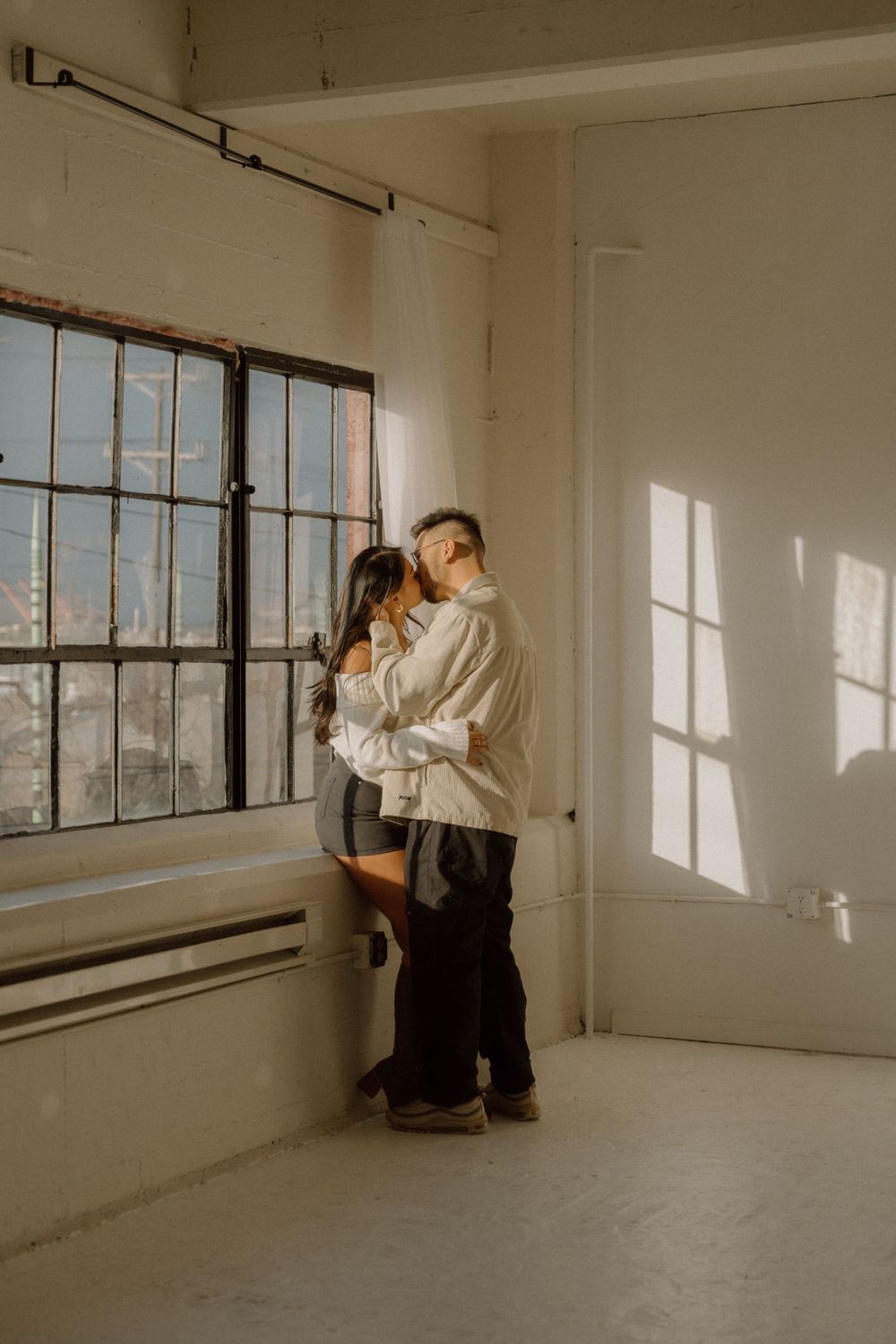 Romantic couple embracing by industrial window in bright minimalist room.