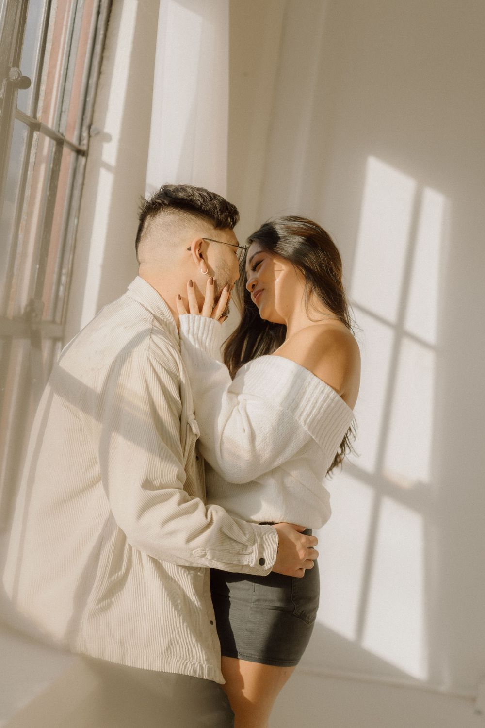 Intimate couple sharing tender moment near window with sun casting dramatic shadows.