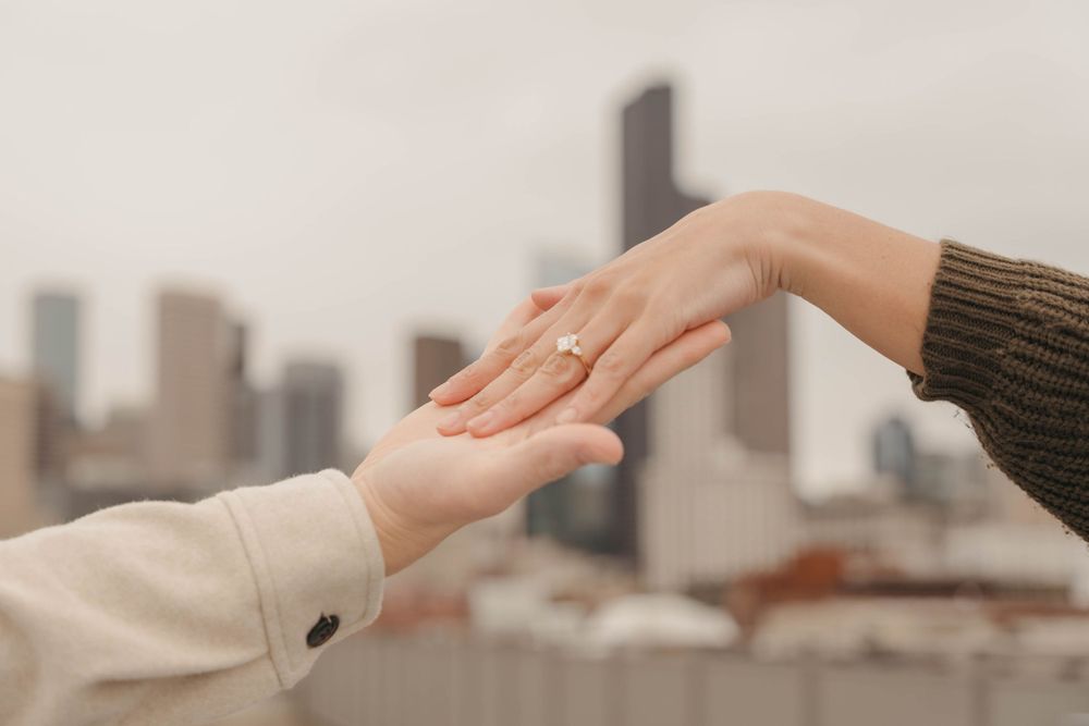 Two hands reaching toward each other with an engagement ring against a city skyline backdrop.