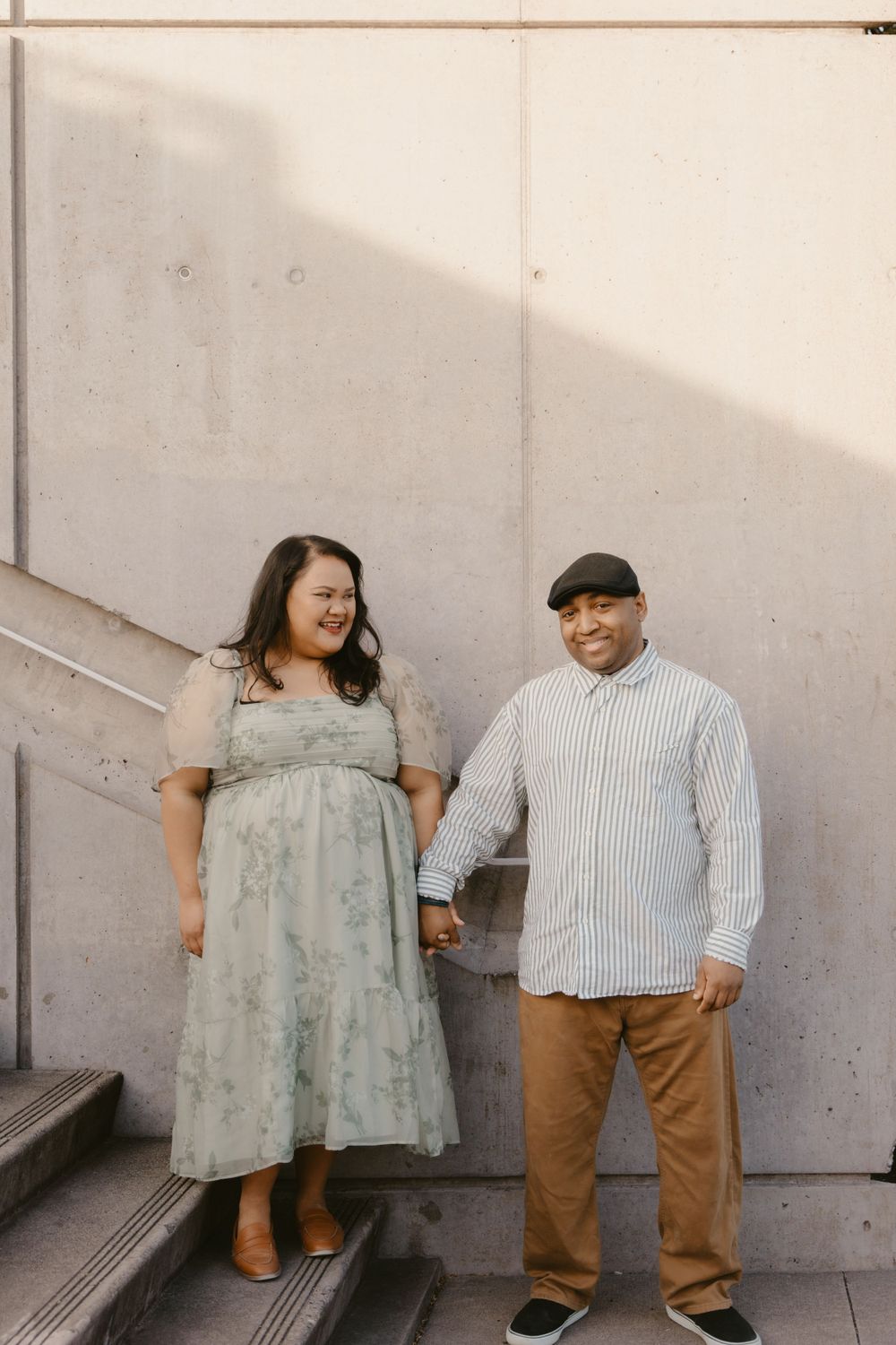 Couple holding hands while standing on concrete steps against white wall.