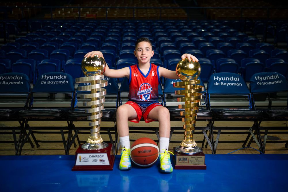 Young athlete posing with basketball and two large golden trophies in stadium.