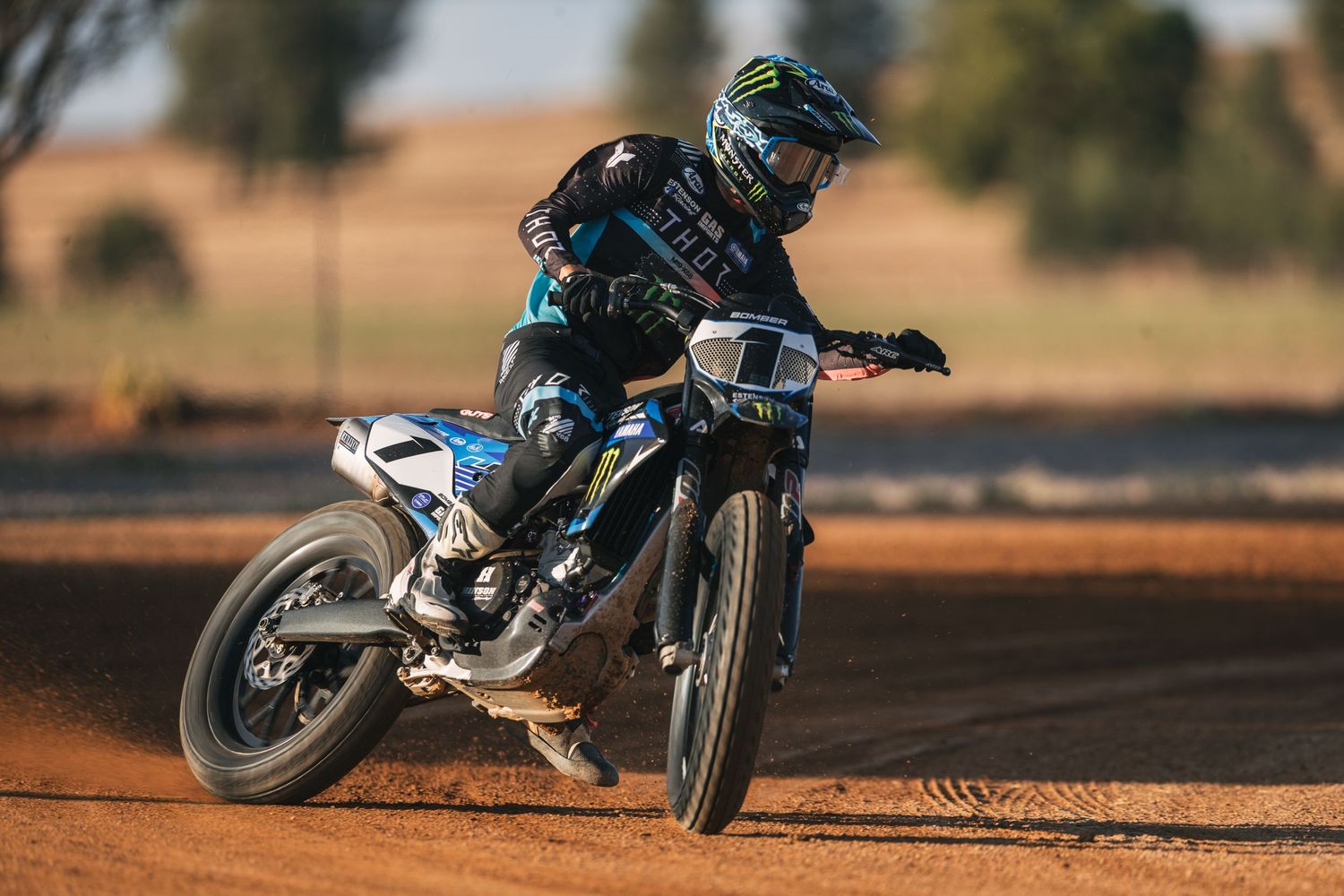 A motocross rider in Monster Energy gear races on a dirt track, leaning into a turn on a blue Yamaha motorcycle.