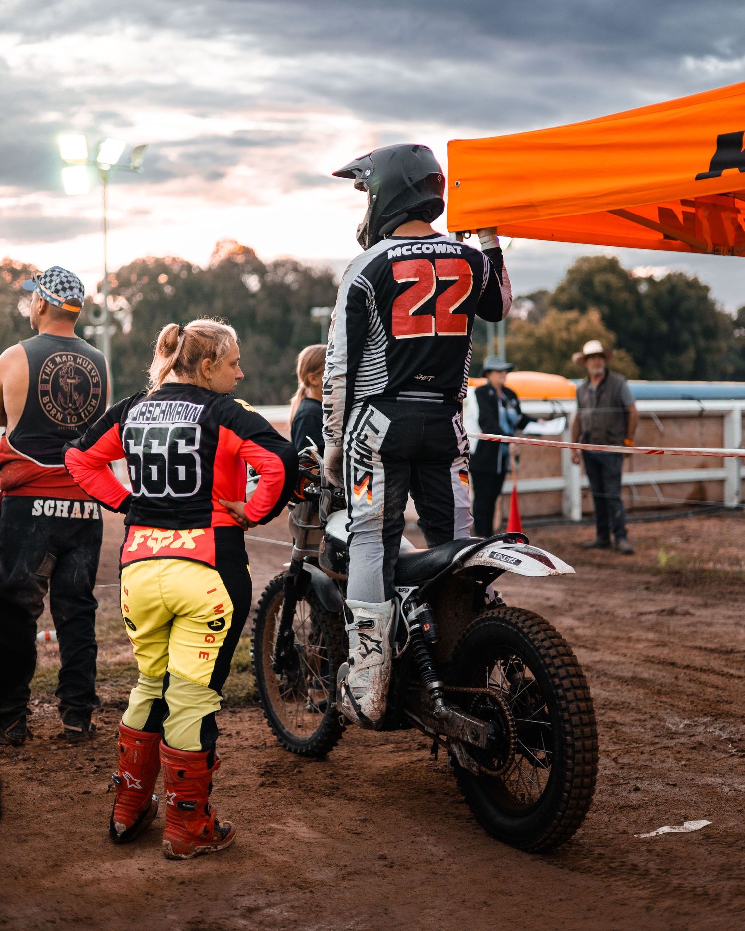 Motocross riders in racing gear standing by their bikes at a dirt track event.