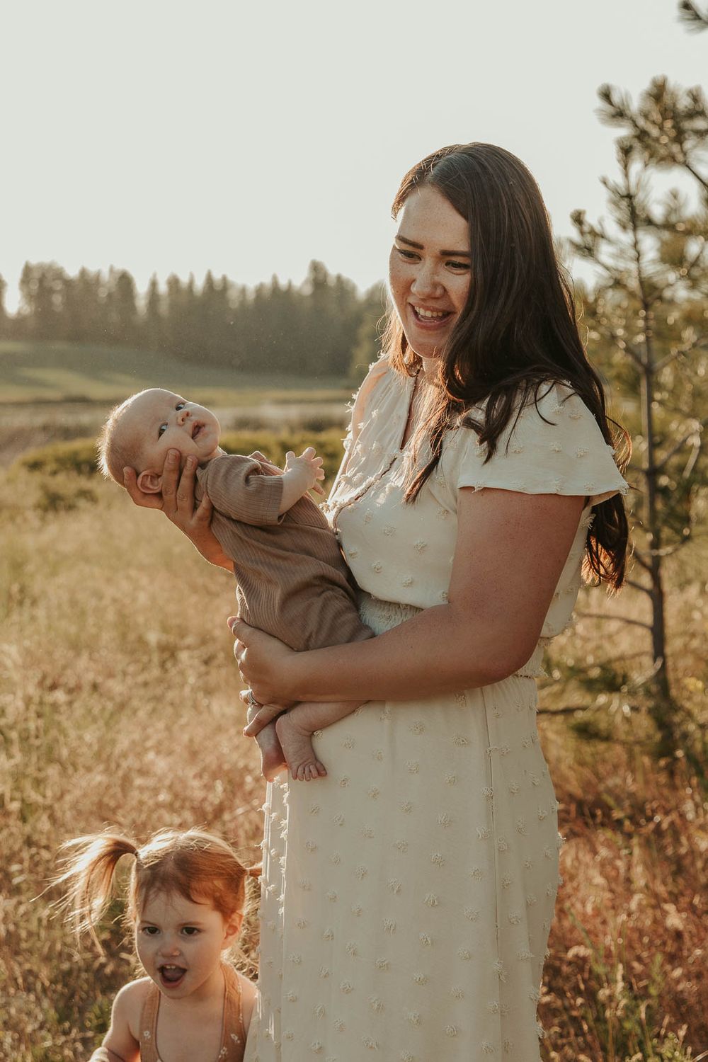 mom with dark hair wearing a white dress smiling and holding a small baby in brown in a field in spokane