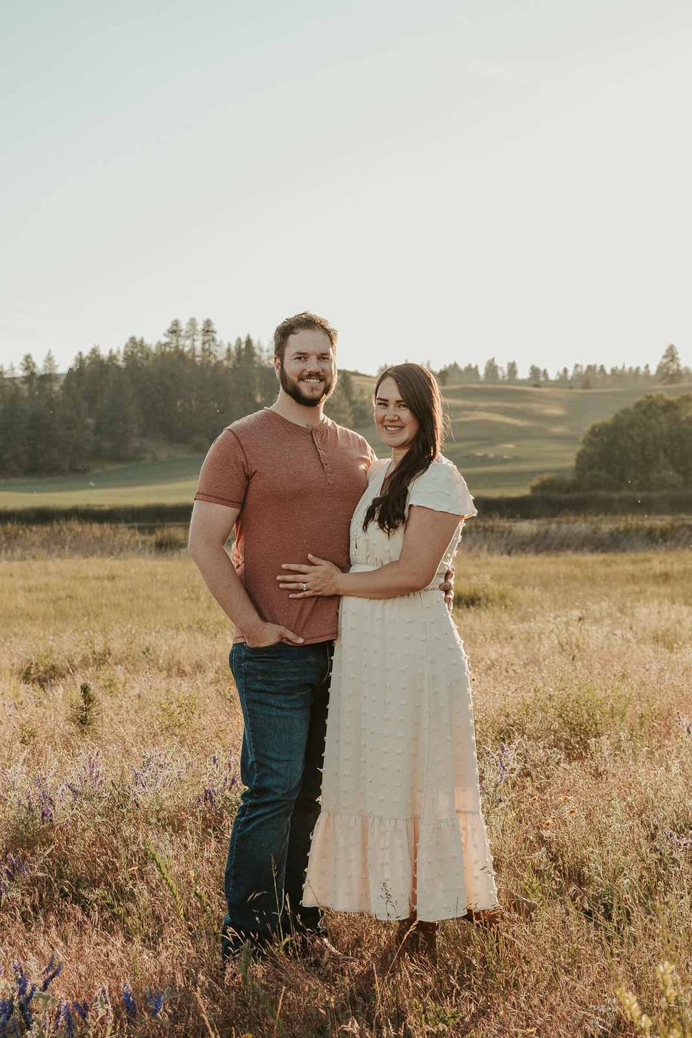 man dressed in brown shirt with woman in white dress smiling at the camera in a spokane open wildflower field