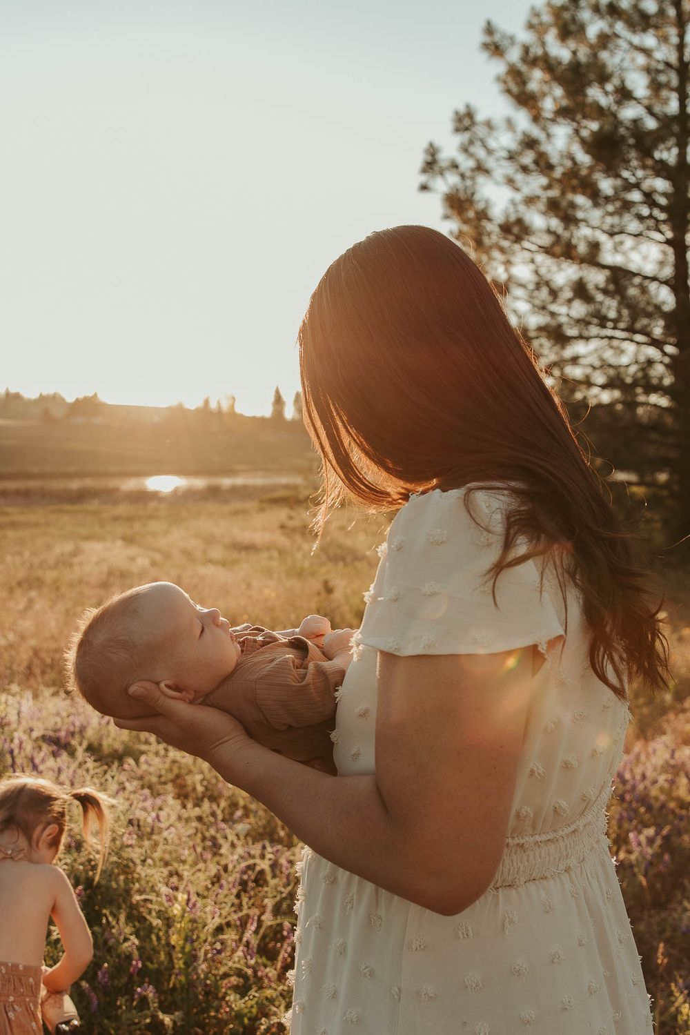 mom with dark hair wearing a white dress smiling and holding a small baby in brown in a field in spokane golden hour
