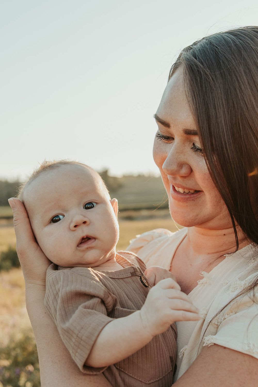mom with dark hair wearing a white dress smiling and holding a small baby in brown in a field in spokane golden hour