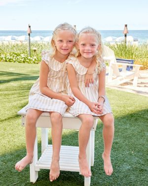 Two young blonde girls in matching striped dresses sitting together on a white bench by the ocean.