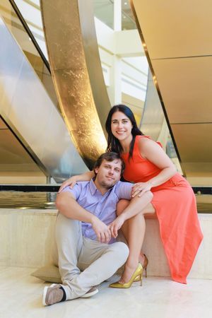 A smiling couple poses in a modern lobby, woman in red dress with gold heels sits above man in casual attire on marble floor.