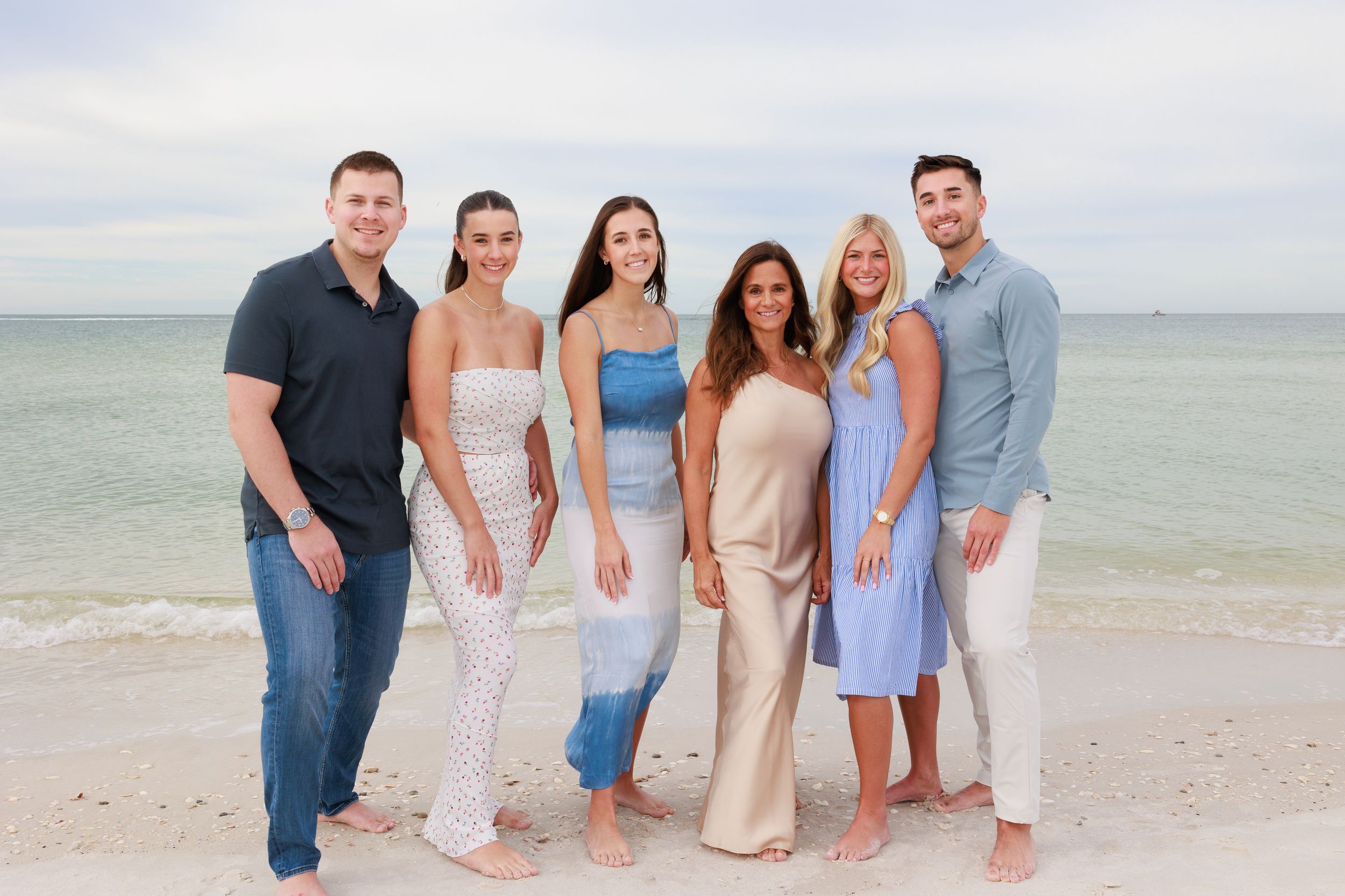 Six people stand together smiling on a sunny beach with calm ocean water behind them.