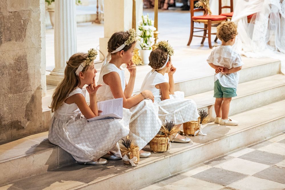 Young attendants in white dresses and flower crowns sit on stone steps.