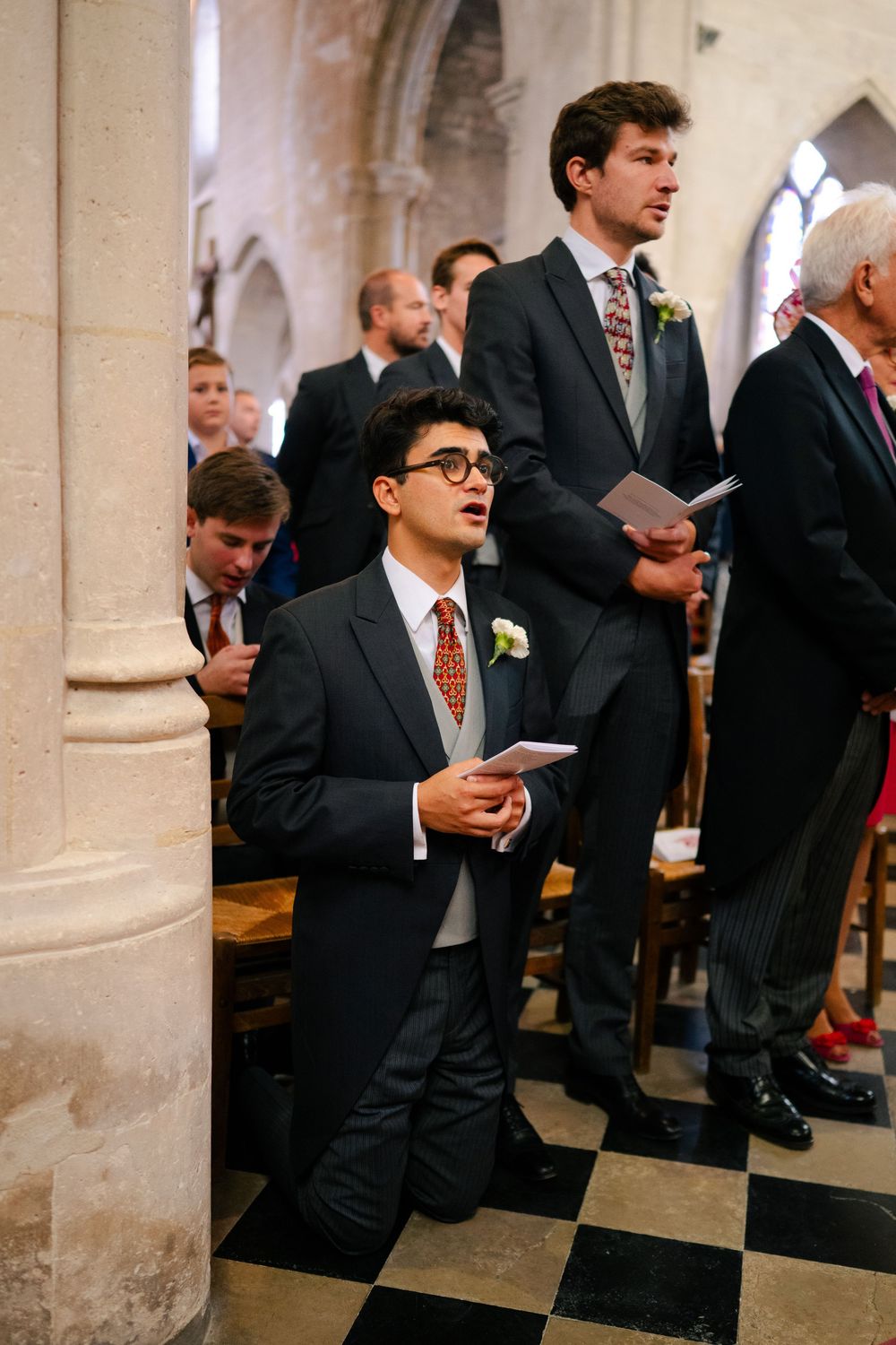 Wedding guests in formal attire stand and sit in church pews during a ceremony.