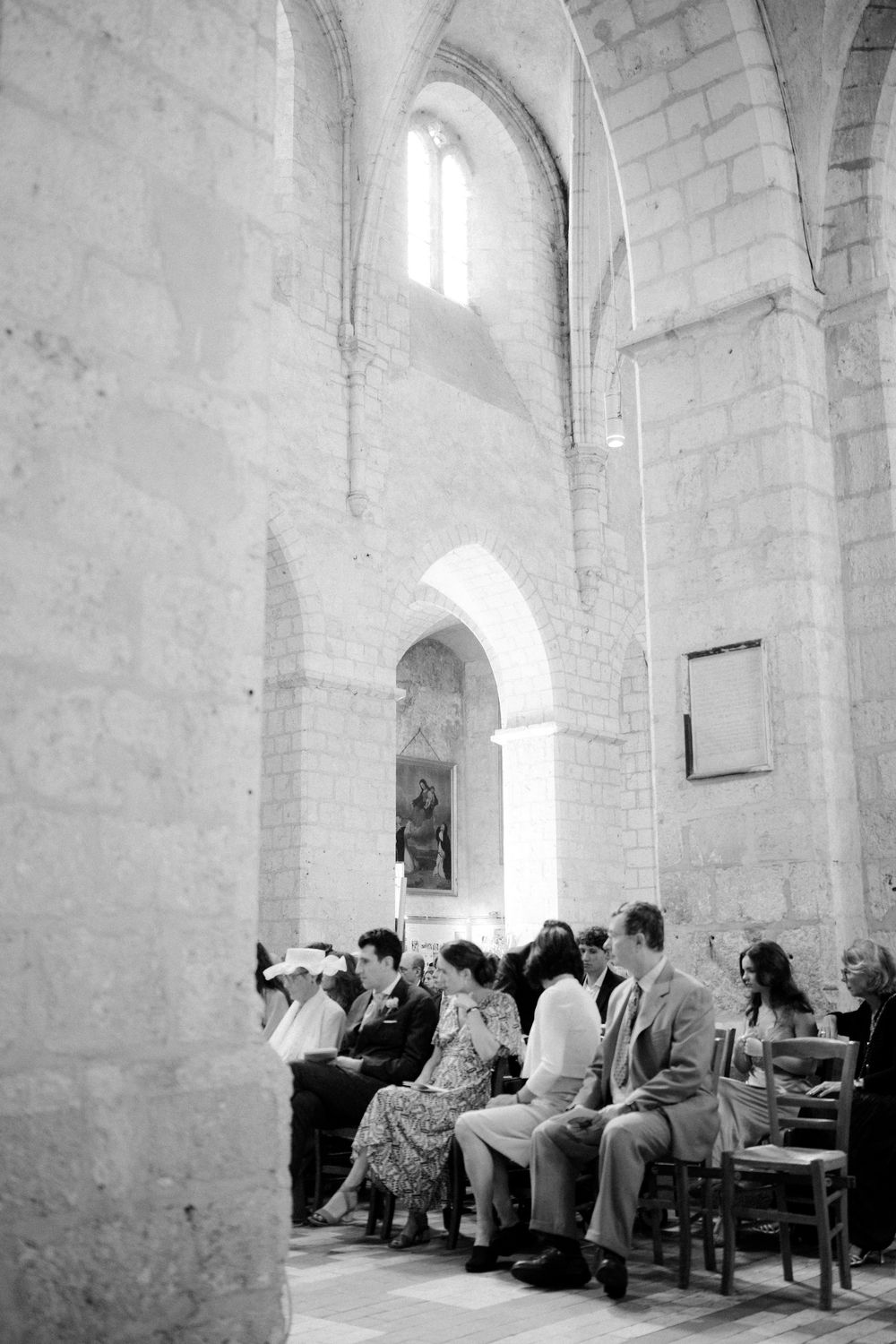 Black and white photo of wedding guests seated in rows inside a historic stone church with arched windows.