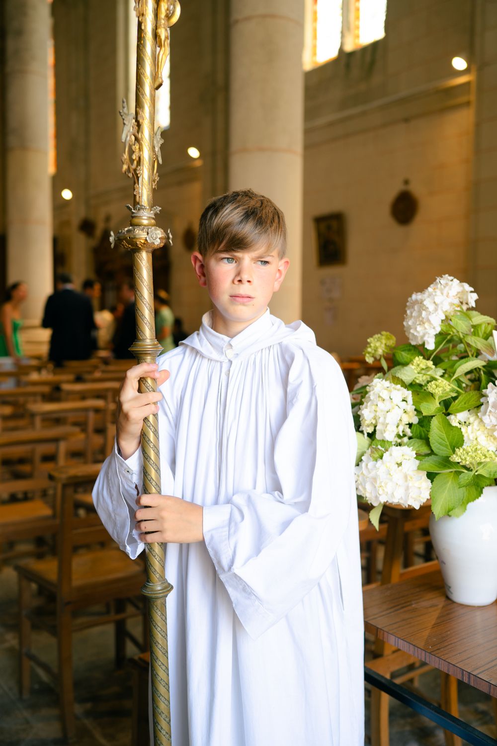 A child in a white choir robe stands in a church interior with white hydrangea flower arrangements and wooden pews.