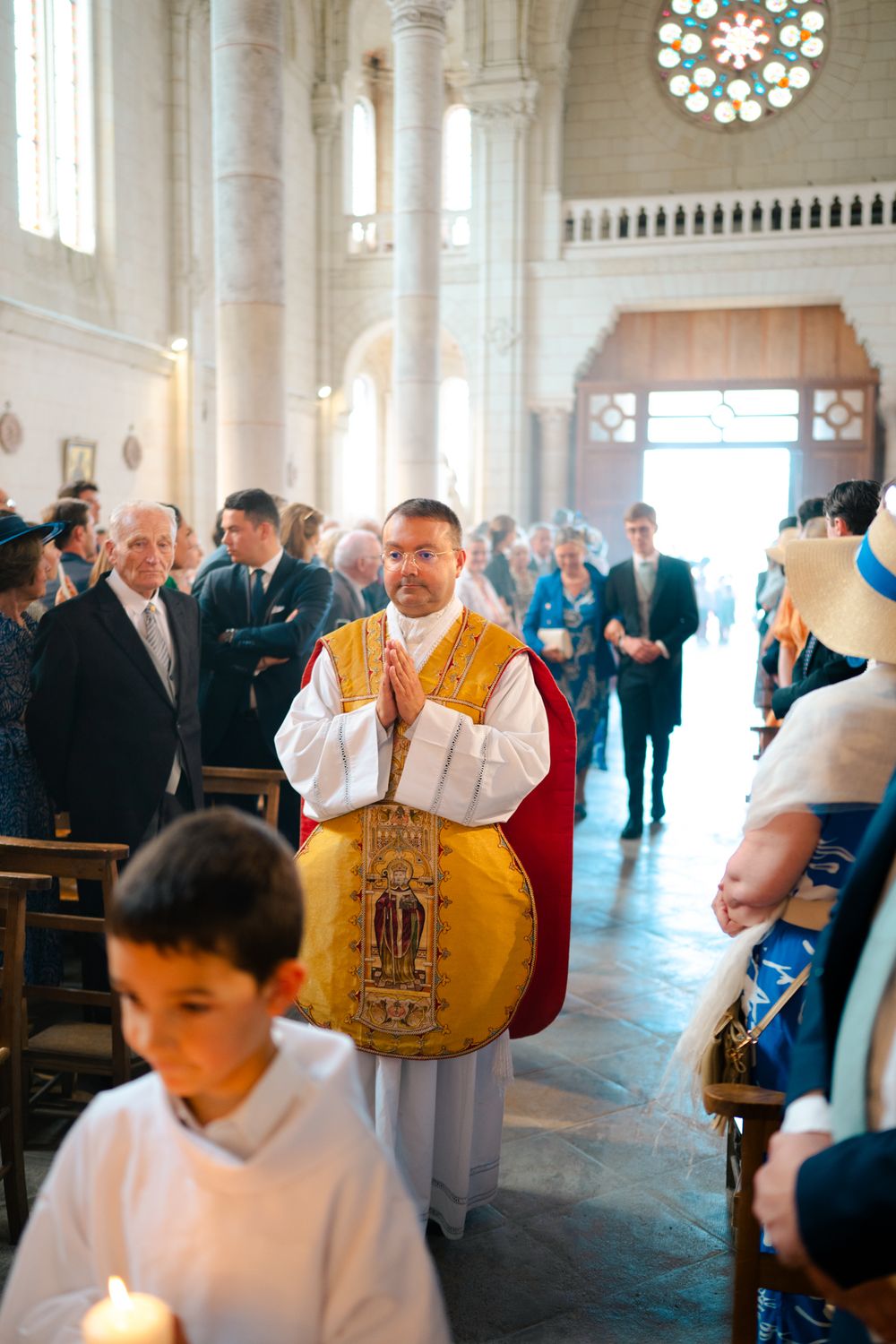 A priest in golden vestments stands in a church aisle during a religious ceremony with a stained glass window visible.