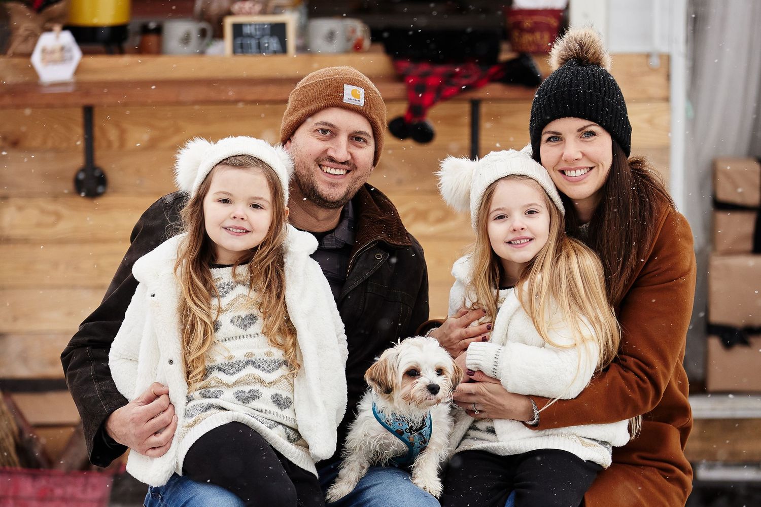 A happy family sits together on a bench with their small white dog for an outdoor winter photo session with a hot chocolate bar behind them.