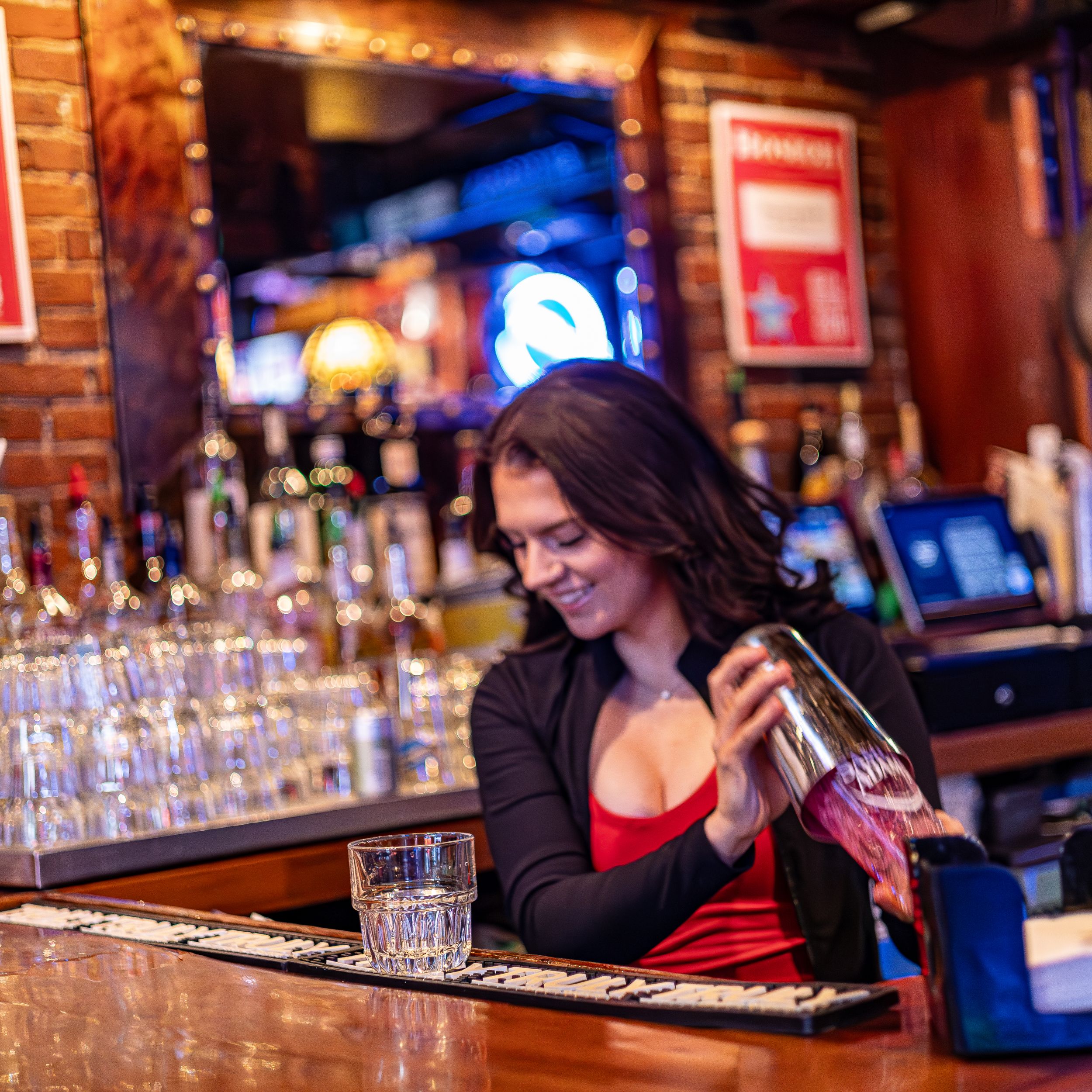 Bartender pouring drinks at a dimly lit bar with brick walls and neon signs in the background.
