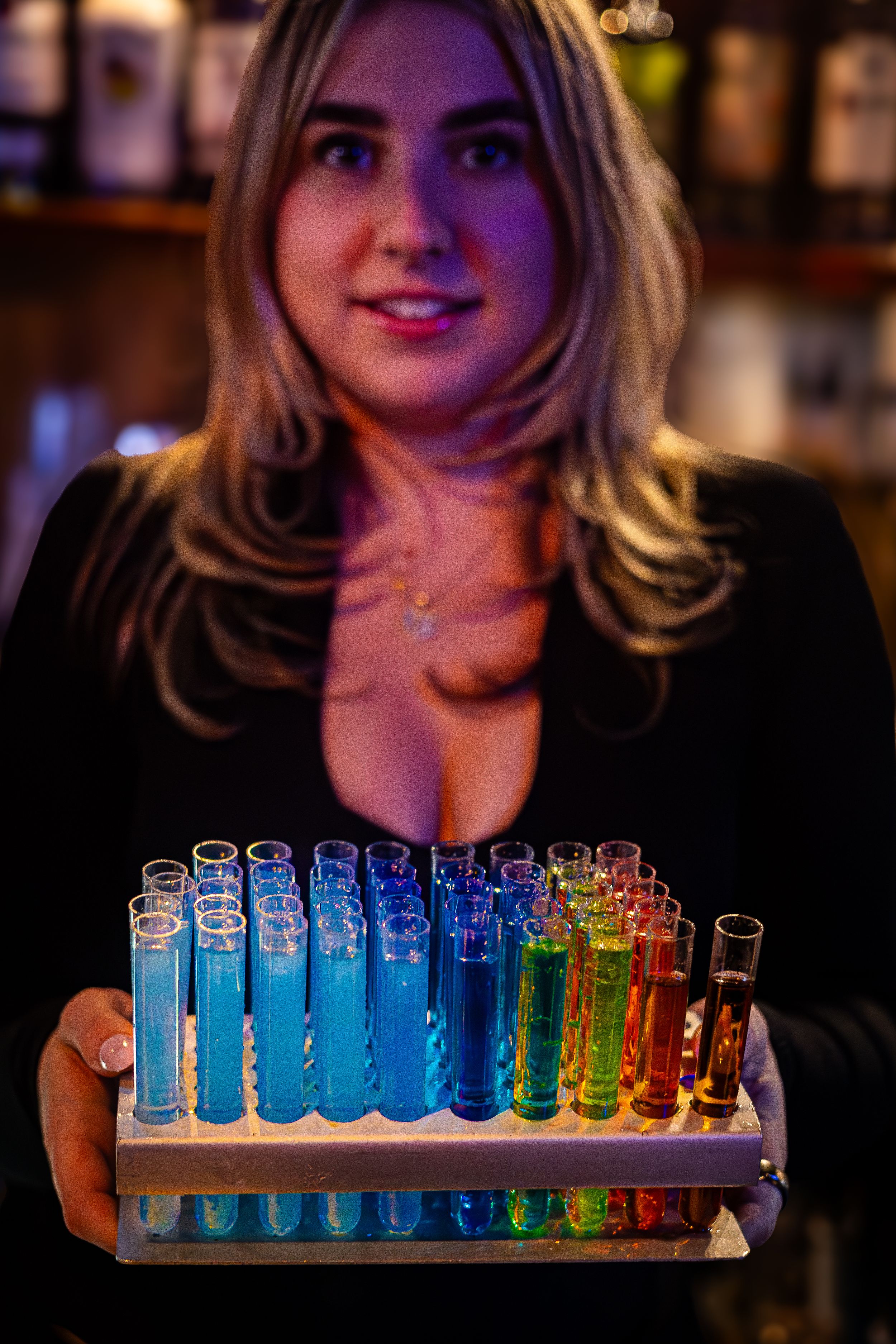 Colorful rainbow test tubes filled with vibrant liquids arranged in a holder being presented at a bar.