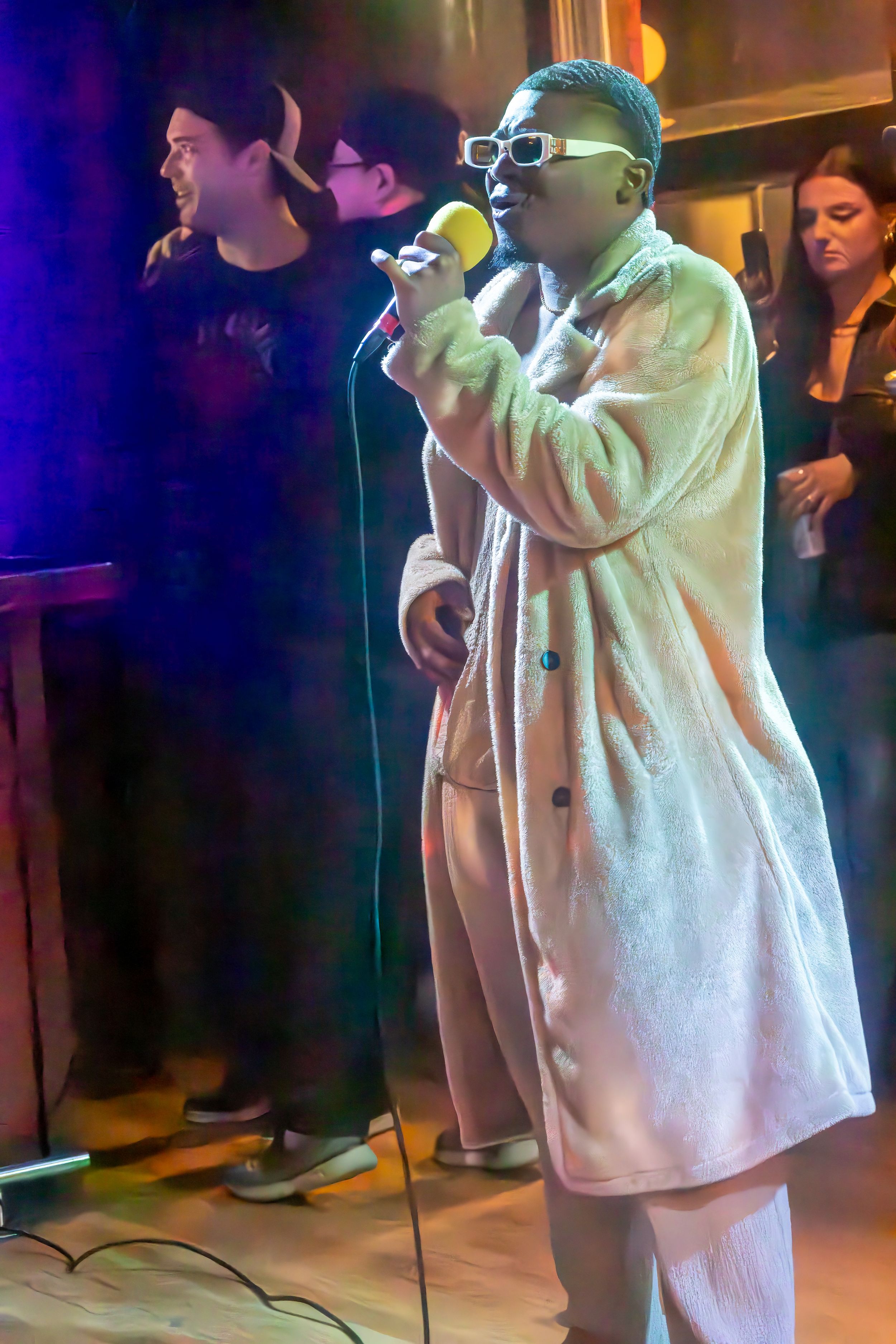 Performer in white coat singing on stage under blue and purple lighting.