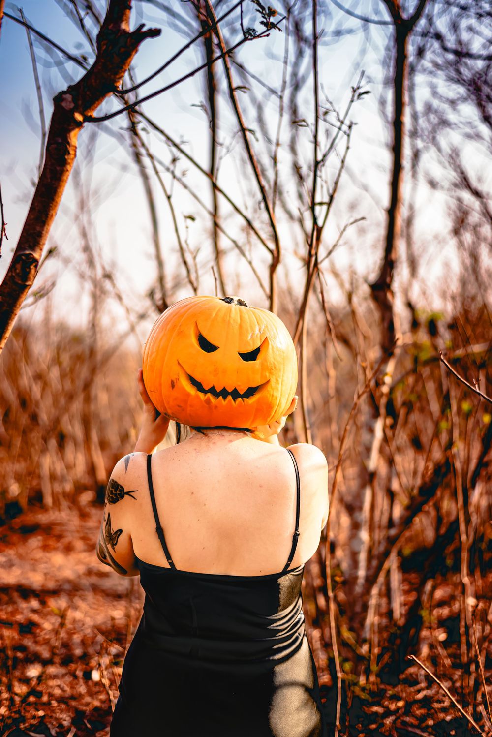 Person wearing black dress holds carved pumpkin on shoulders in autumn nature setting.