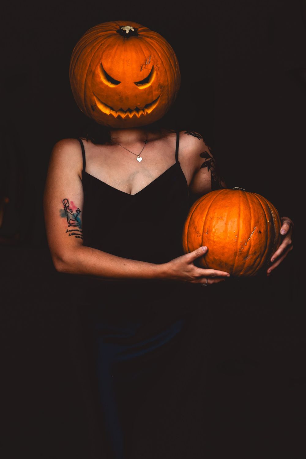 Creative Halloween portrait with carved pumpkin head and person holding another pumpkin against dark background.