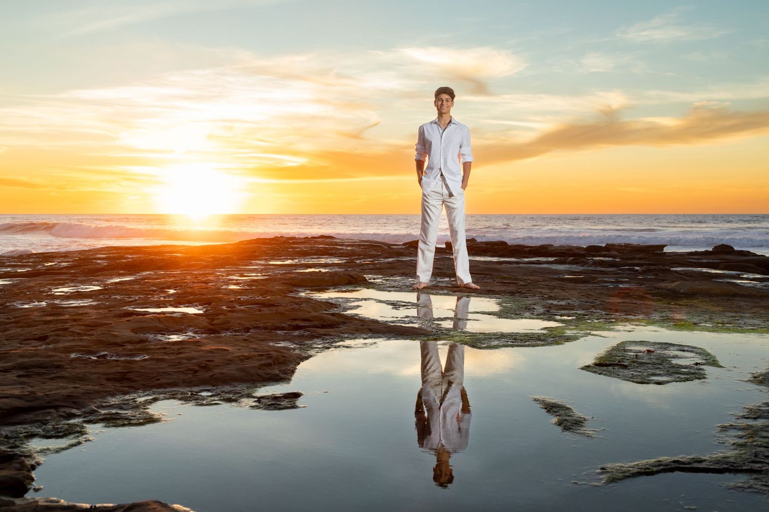 A high school senior in white clothing stands on a rocky beach at sunset with their reflection mirrored in a tidal pool.