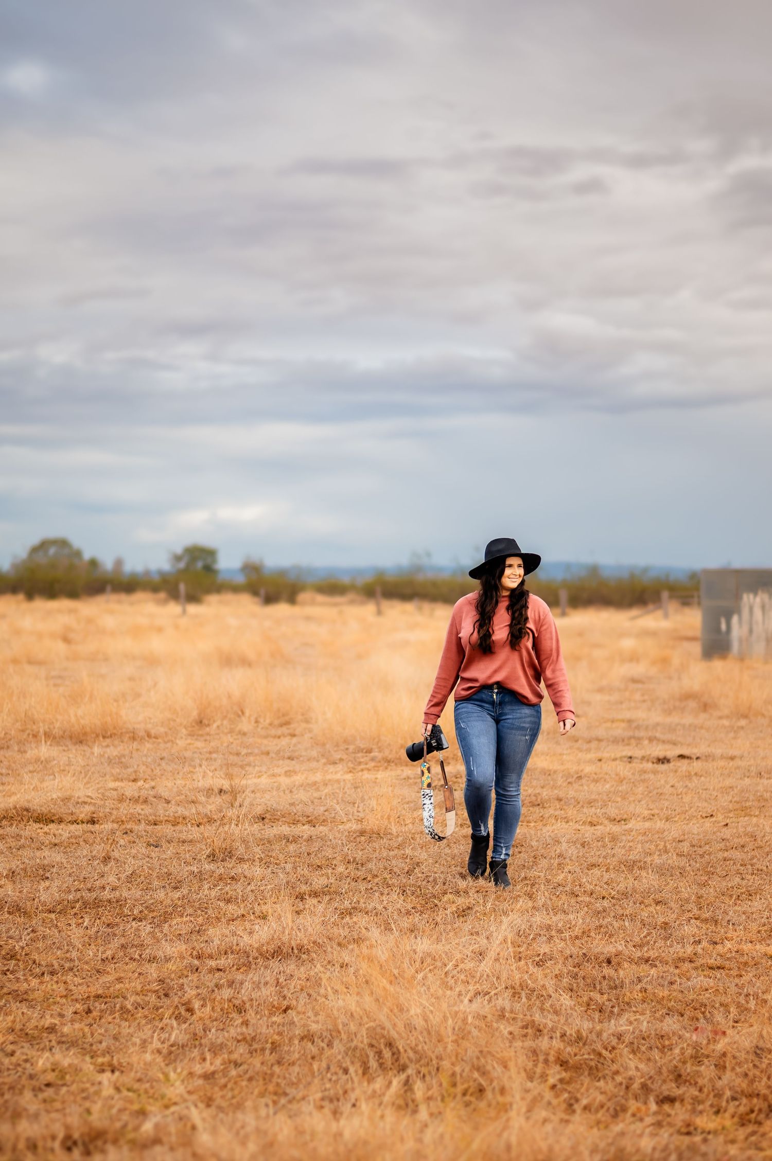 Photographer in casual attire walking through dry grass field with camera gear.