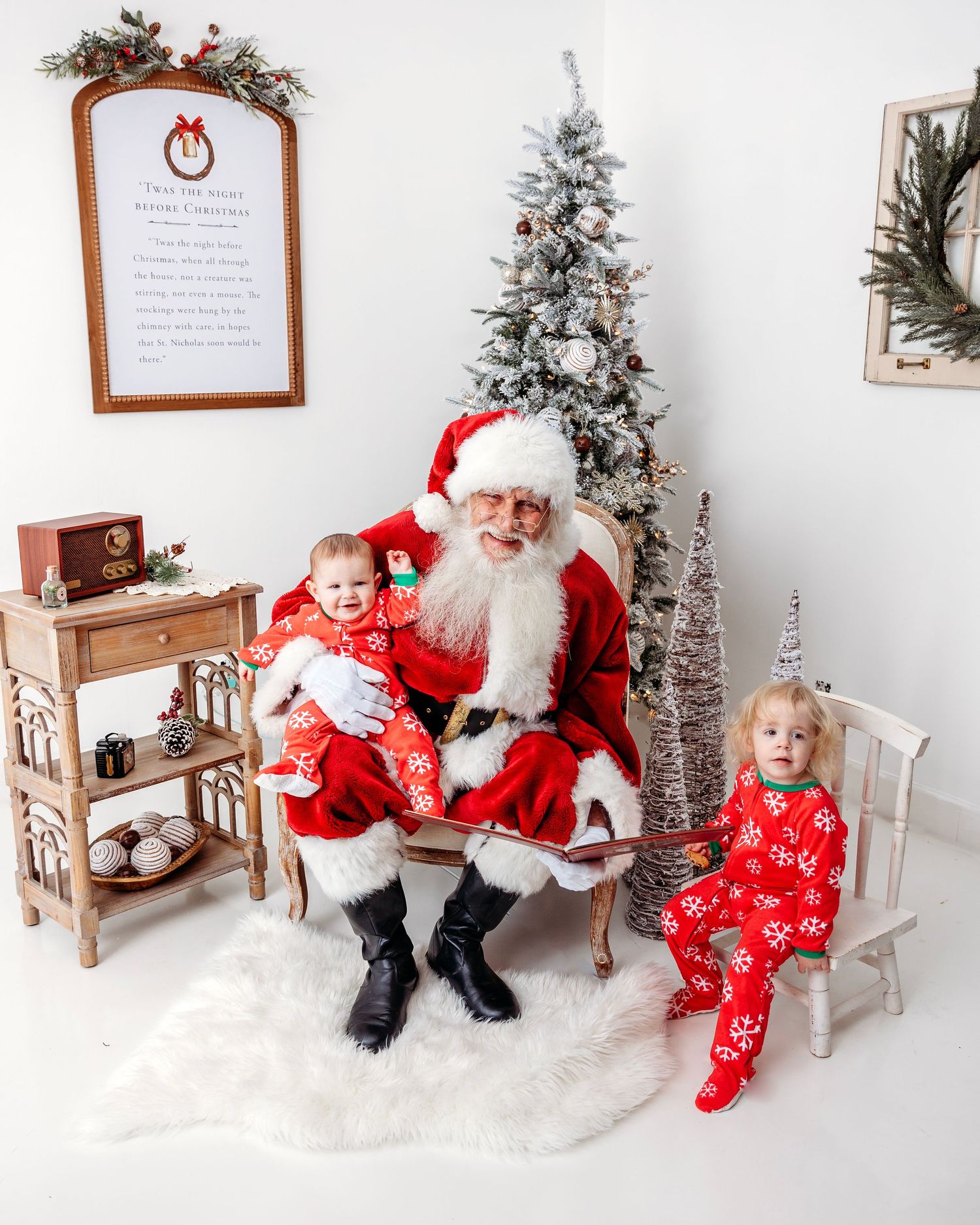 Children in matching red pajamas visit with Santa Claus in a cozy white studio setting with Christmas decorations.
