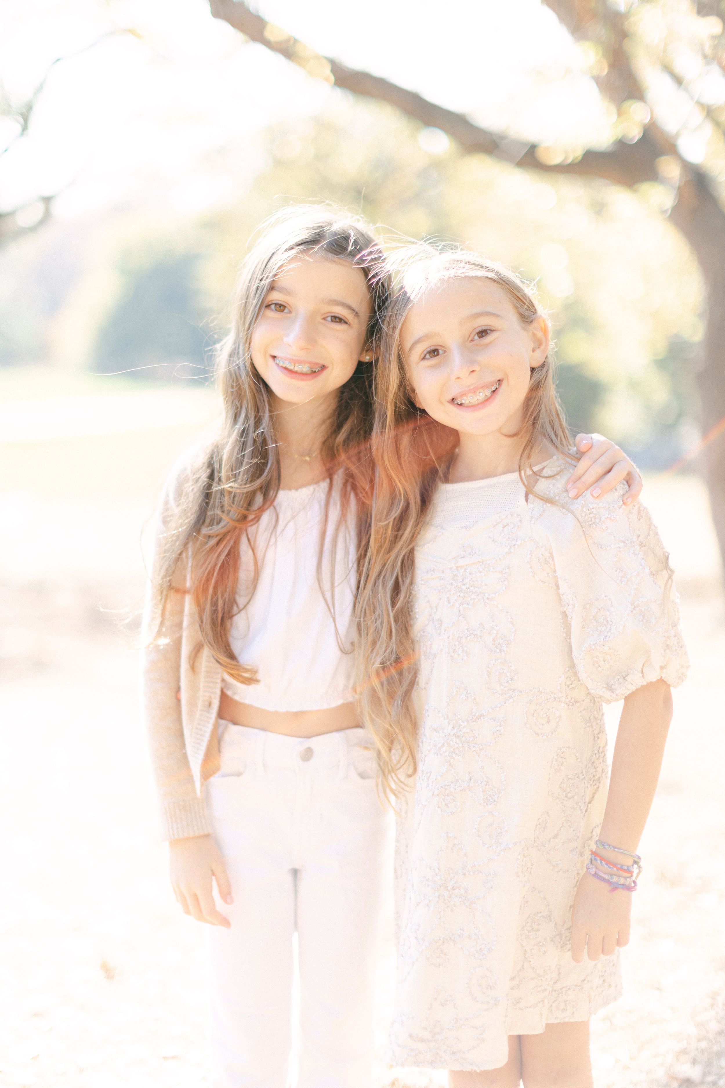 Two young people in light colored outfits share a friendly moment outdoors in soft sunlight.