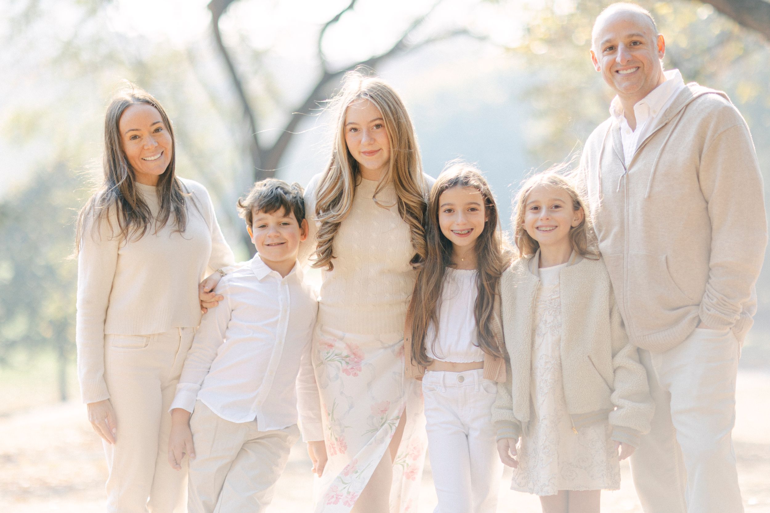 Family group in coordinated neutral tones poses together in a sunlit outdoor setting.