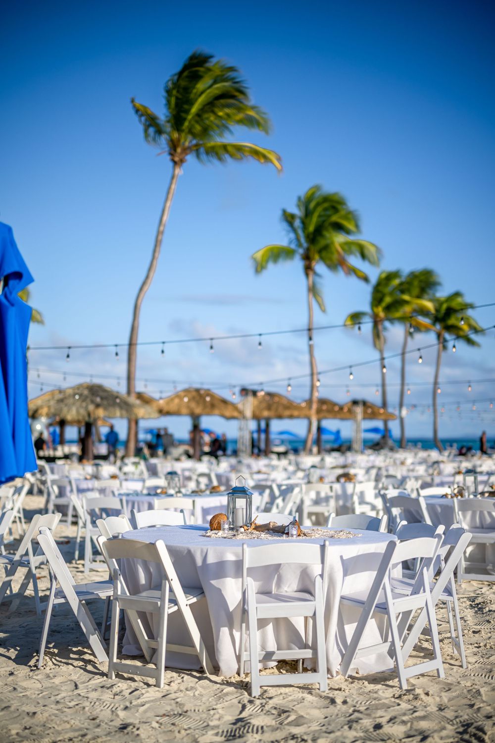 White tables and chairs set up for a beach wedding reception with palm trees and tiki umbrellas in the background.