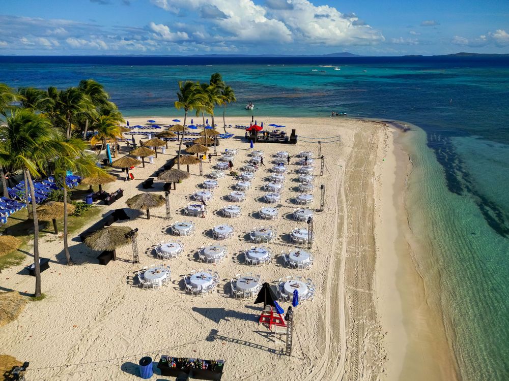 Aerial view of beach resort dining setup with white tables and umbrellas along turquoise shoreline.