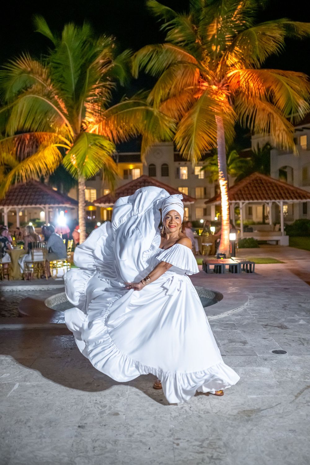 Dancer in flowing white dress performs under palm trees at night with resort lighting.