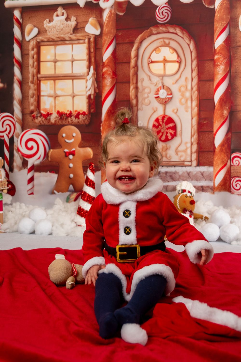 Toddler wearing Santa outfit smiles while sitting on red blanket with gingerbread house backdrop.