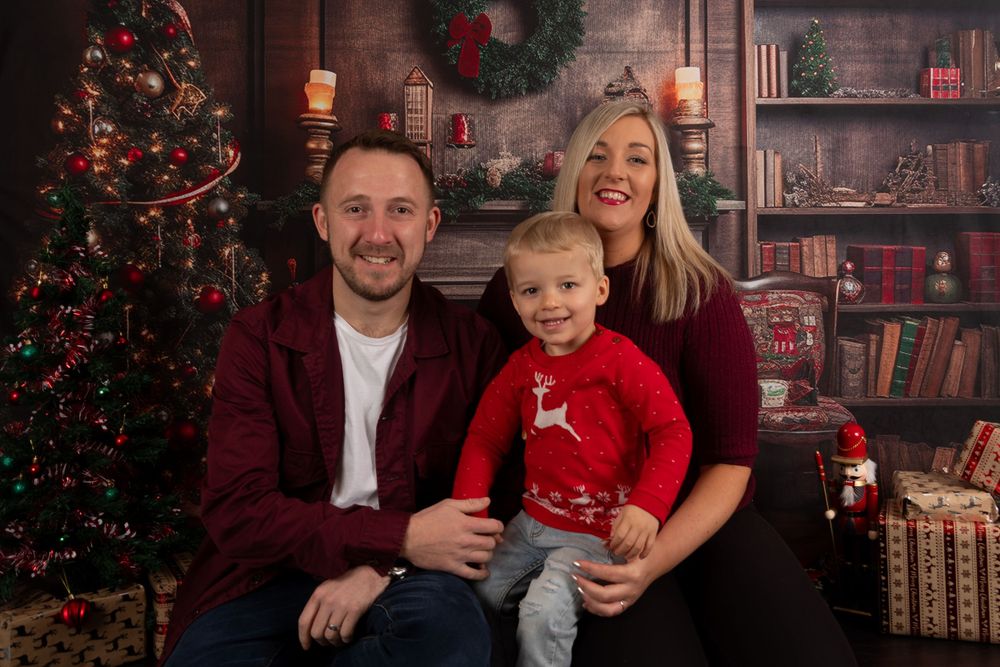 Family of three poses together in front of Christmas tree and decorated fireplace.